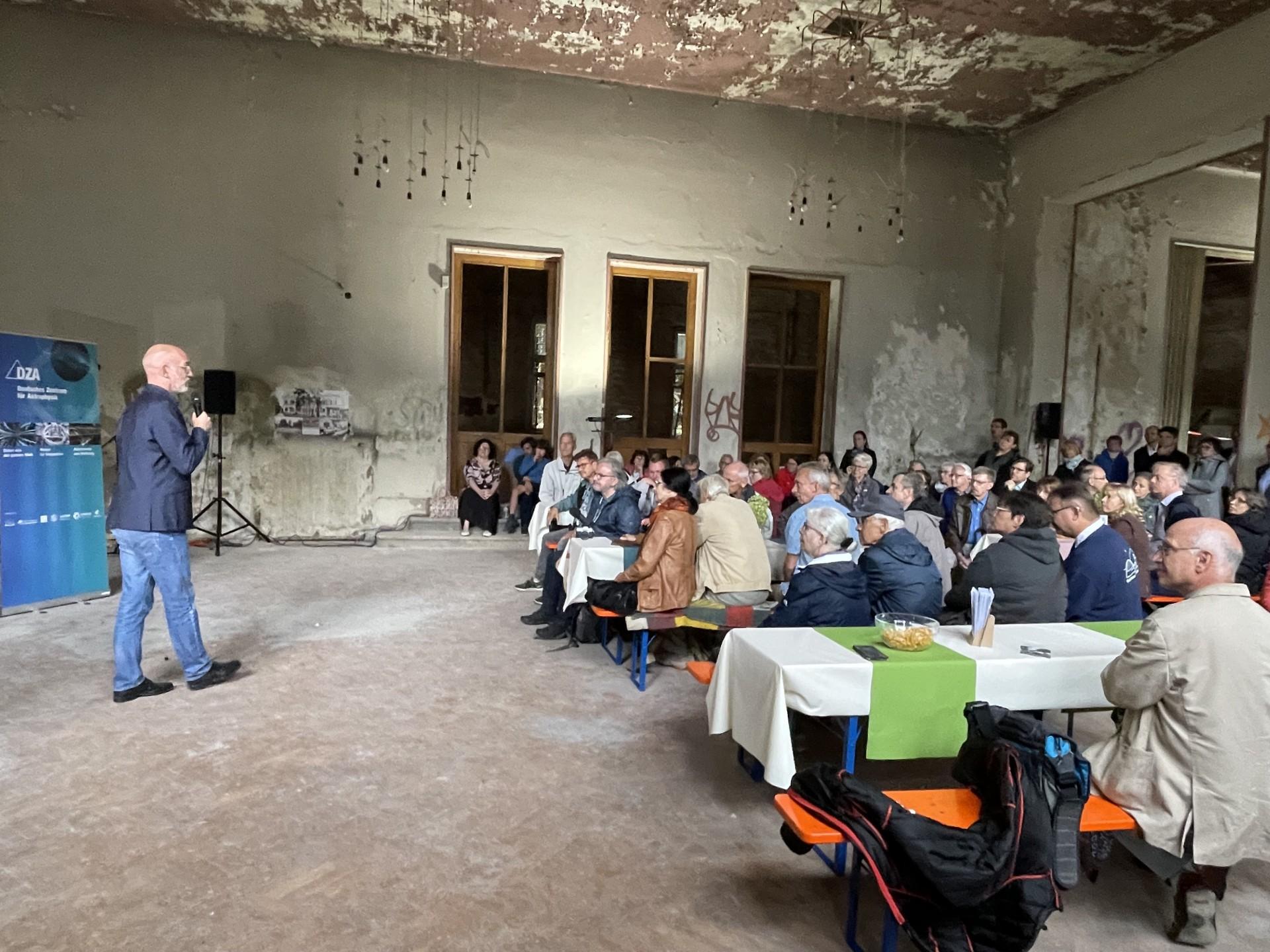 viele Besucher sitzen in einer heruntergekommenen Halle und hören einem Vortragenden zu