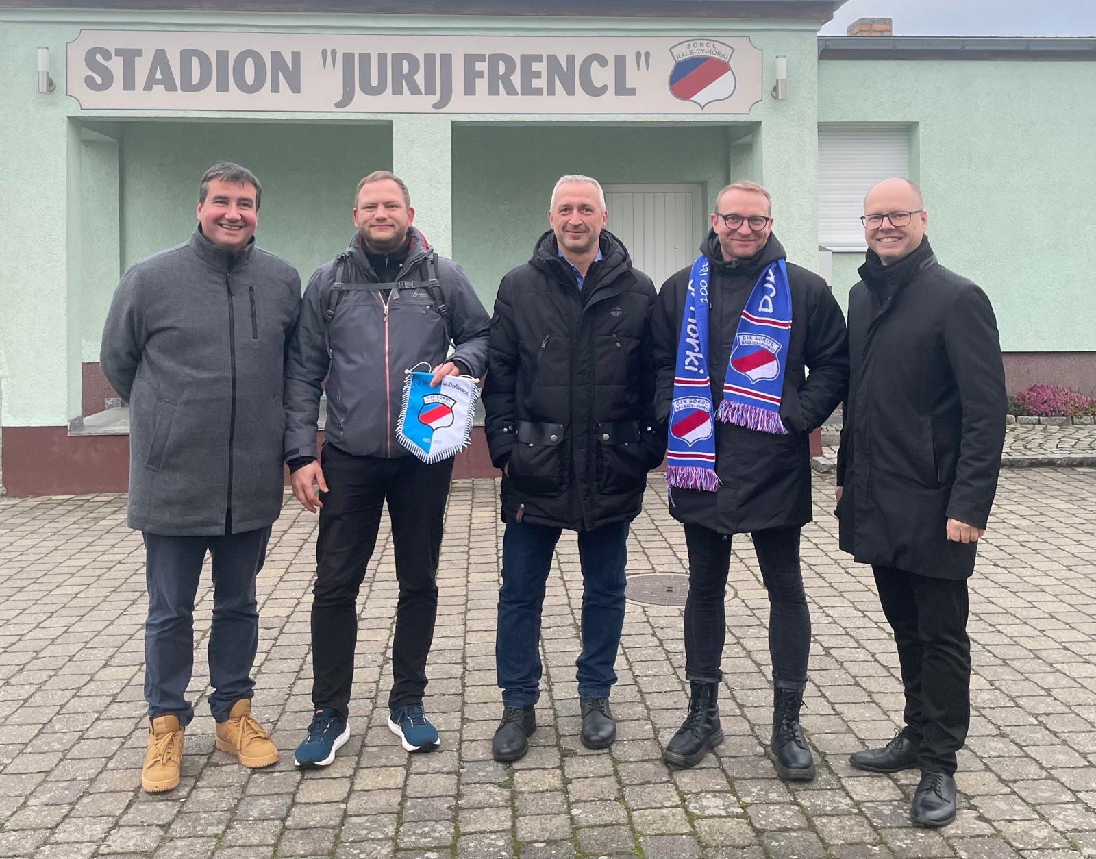 Das DZA mit Dierk Schuster (Präsident DJK), Sebastian Miersch (ehrenamtlicher Jugendtrainer) und Dawid Statnik (Vorsitzender der Domowina) fünf Personen stehen vor einem Stadion mit der Aufschrift "Jurij Frencl"