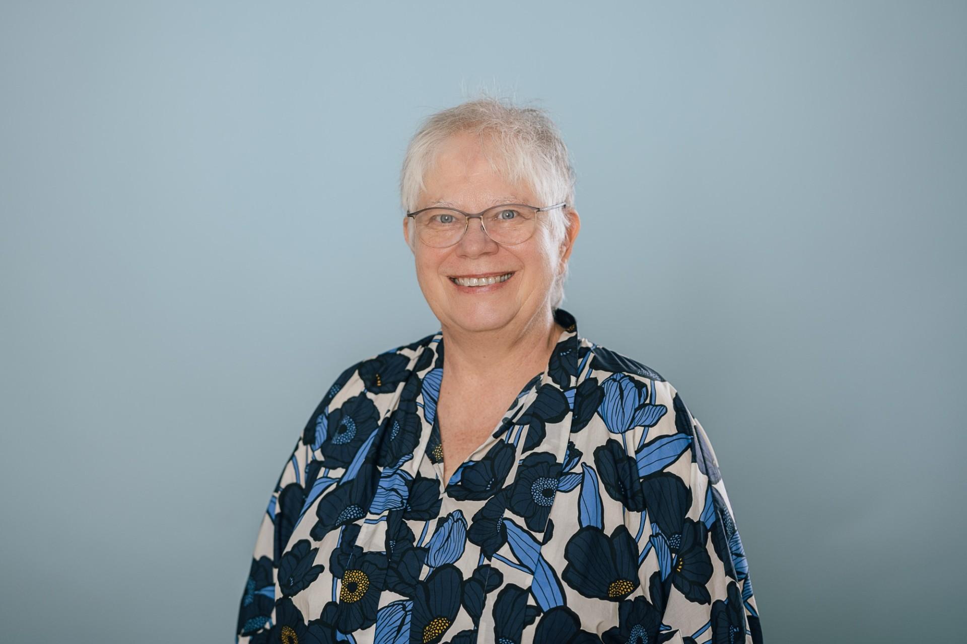Frau mit kurzen weißen Haaren, einer Brille und bunter Bluse lächelt freundlichin Kamera