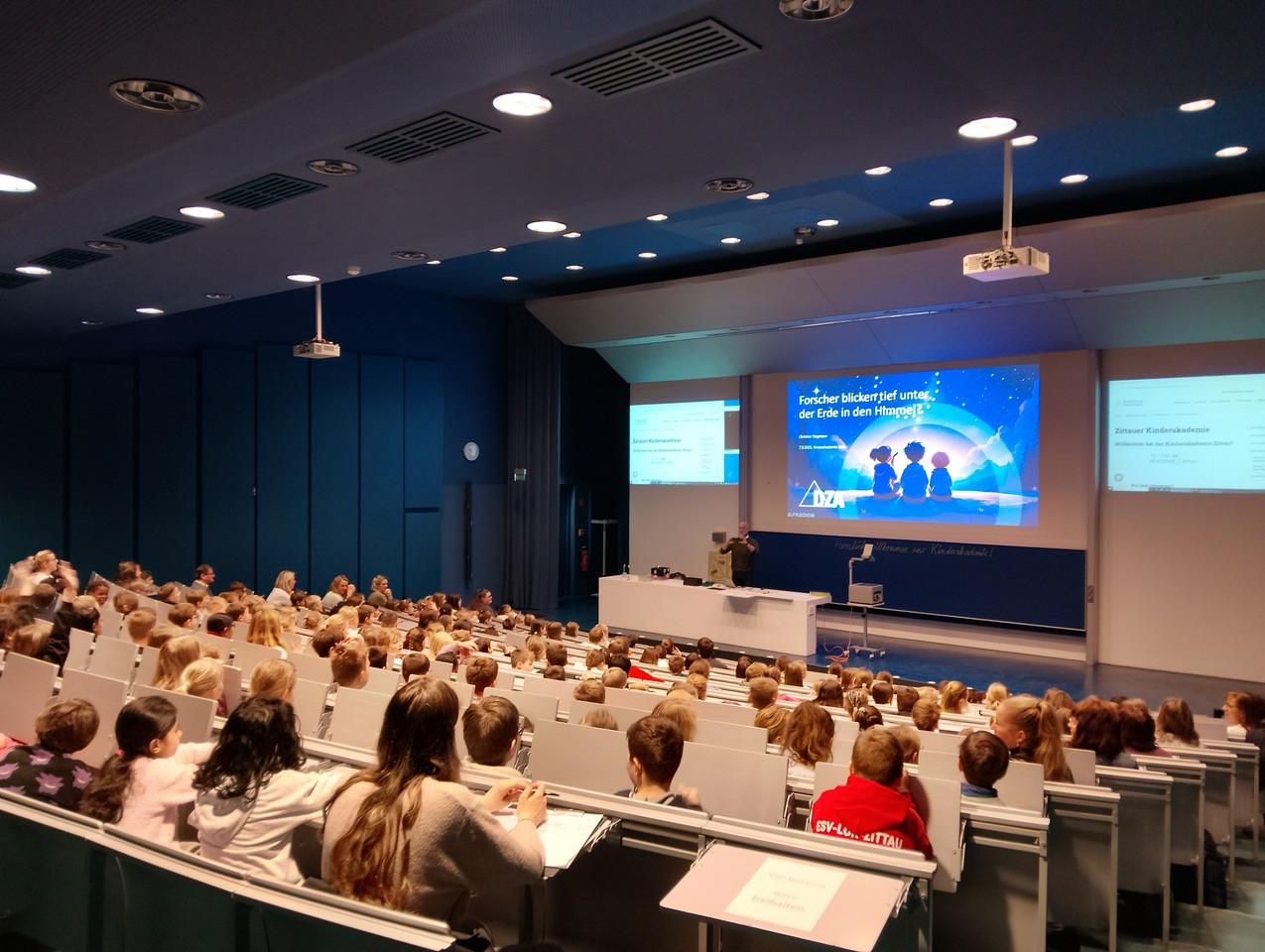 DZA-Vortrag Christian Stegmann auf Kinderakademie Kinder in einem Hörsaal schauen auf einen blauen Screen mit Aufschrift DZA
