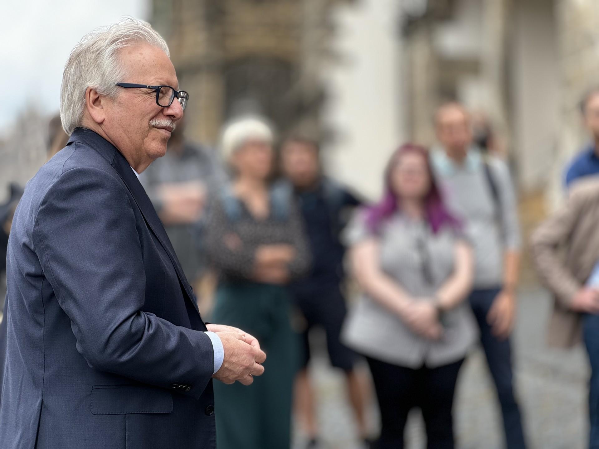 Mann mit Brille und grauen Haaren steht vor unscharfer Menschengruppe im Hintergrund