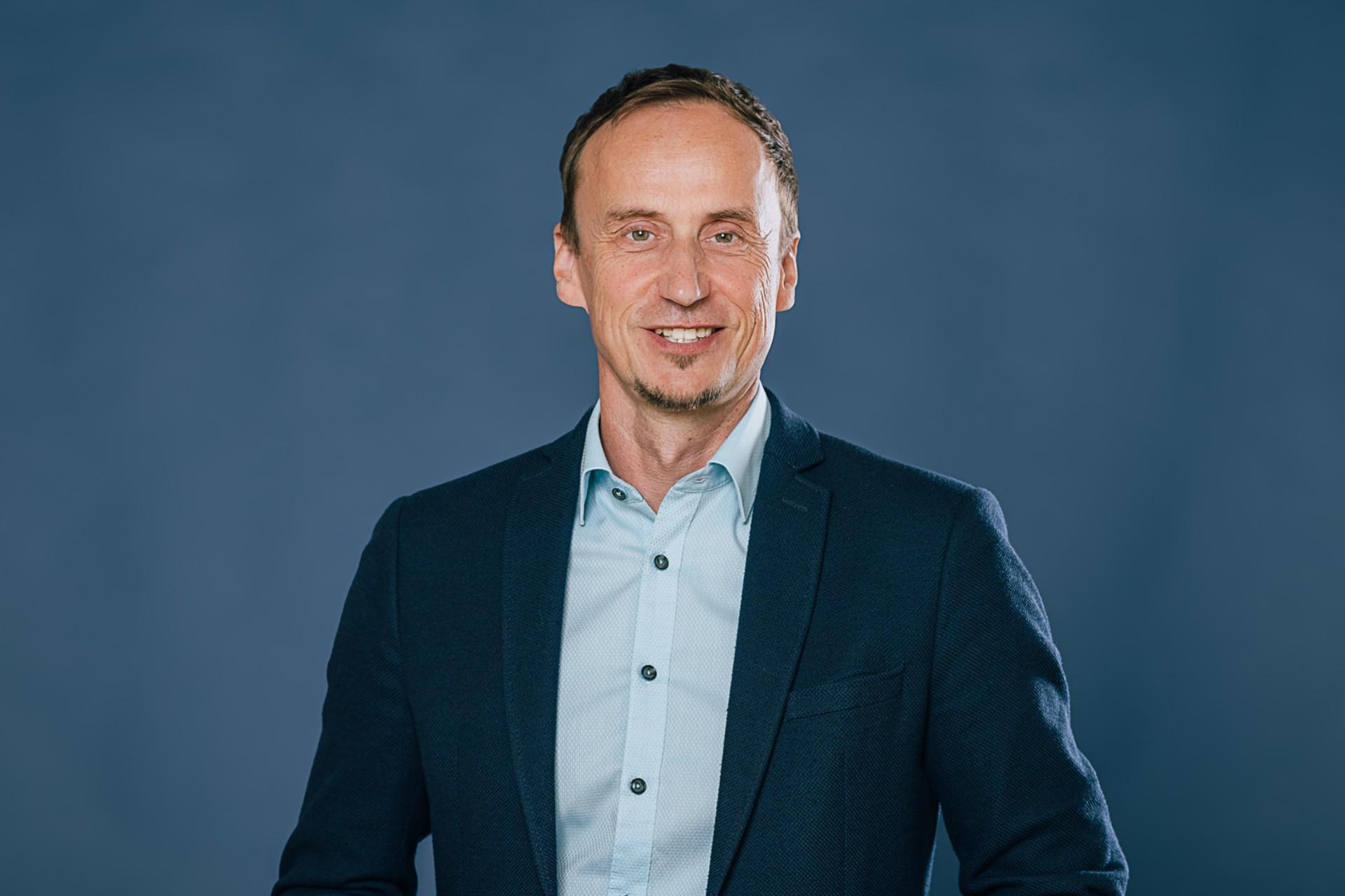 Mann mit lächelndem Gesicht und kurzen dunklen Haaren, trägt ein schwarzes Sakko