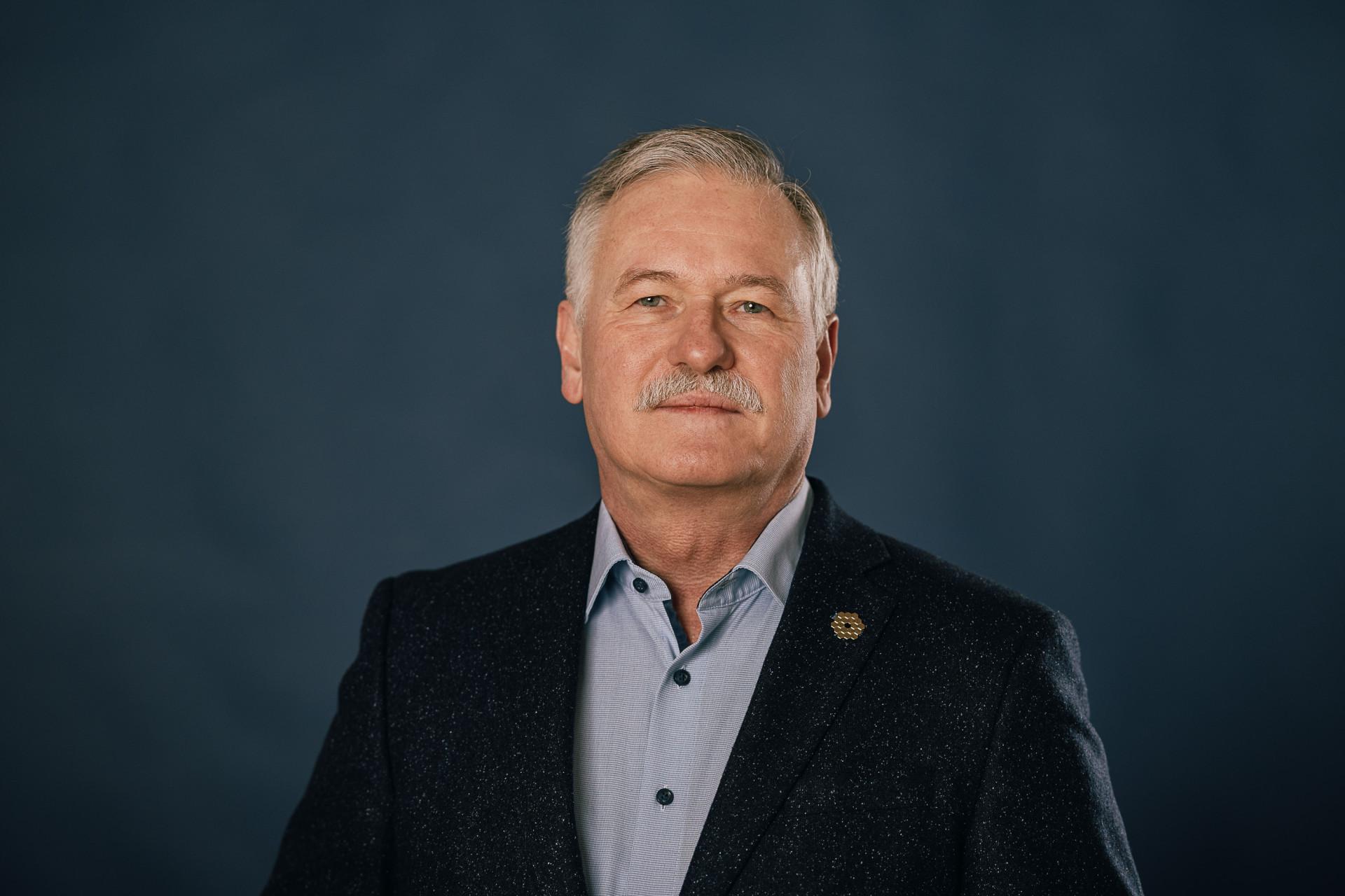 Mann mit Schnauzbart und kurzen Haaren in blauem Hemd und mit Anzug lächelt in Kamera