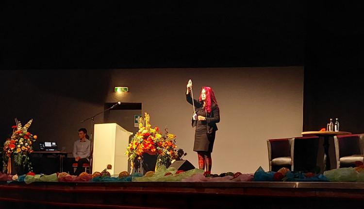 eine Frau mit violetten Haaren steht auf einer Bühne und präsentiert ein Experiment