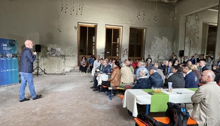 viele Besucher sitzen in einer heruntergekommenen Halle und hören einem Vortragenden zu